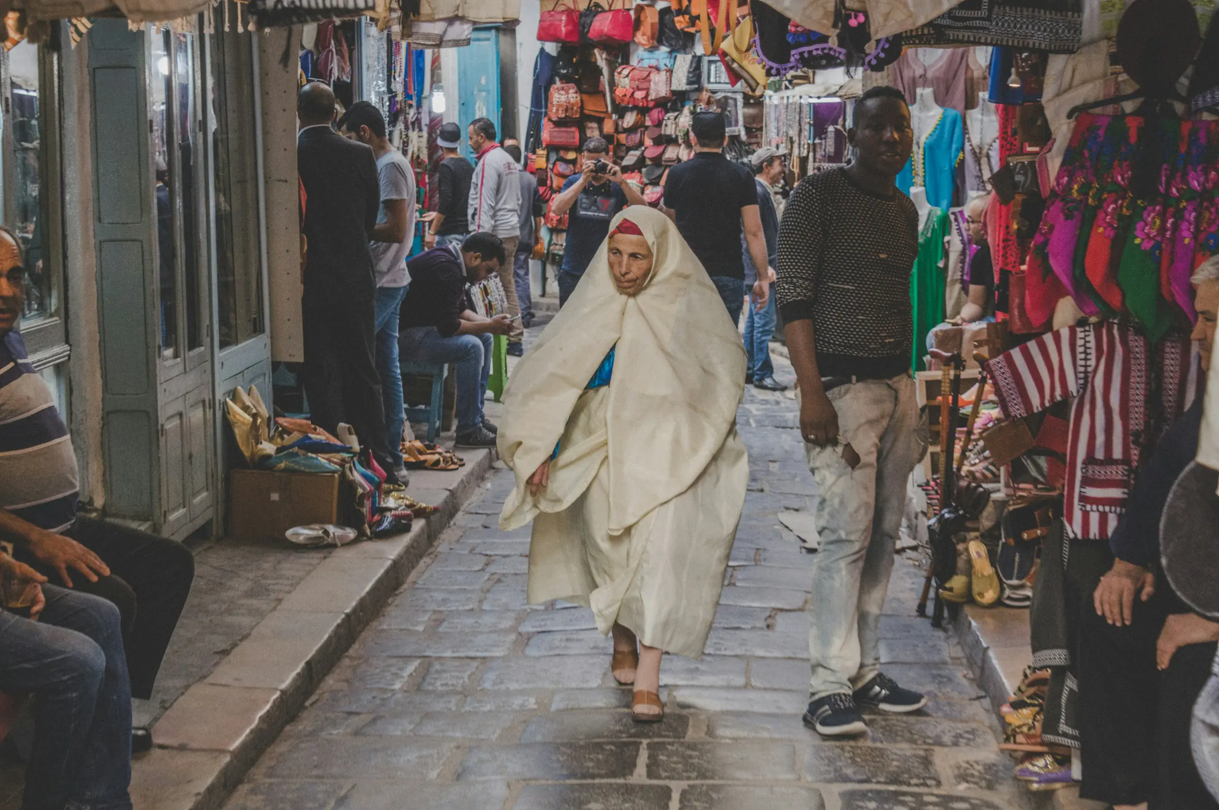 A busy covered souk in the Tunis medina, with shoppers walking between stalls under a vaulted ceiling